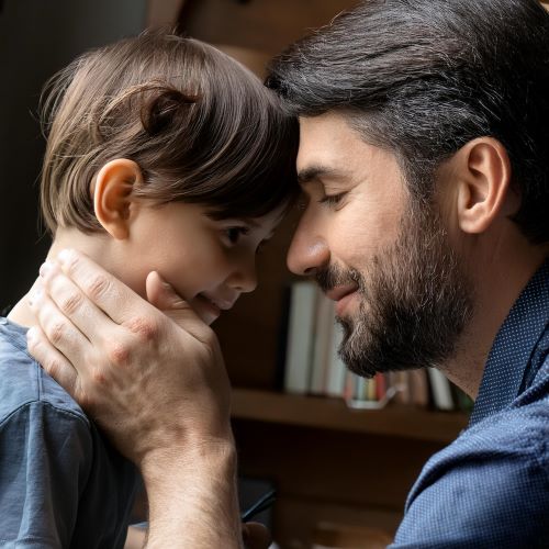 L'immagine mostra un genitore, probabilmente una madre, che si china con espressione calma e comprensiva verso il proprio figlio, un bambino di circa 5-6 anni. La madre tiene delicatamente le mani del bambino mentre lo guarda negli occhi, suggerendo un momento di connessione emotiva e di ascolto attento. Il bambino ricambia lo sguardo con un'espressione di fiducia e apertura. Entrambi sono seduti o accovacciati a terra, indicando un approccio genitoriale basato sull'essere al livello del bambino sia fisicamente che emotivamente. Lo sfondo è sfocato, mettendo in risalto l'interazione intima tra genitore e figlio.